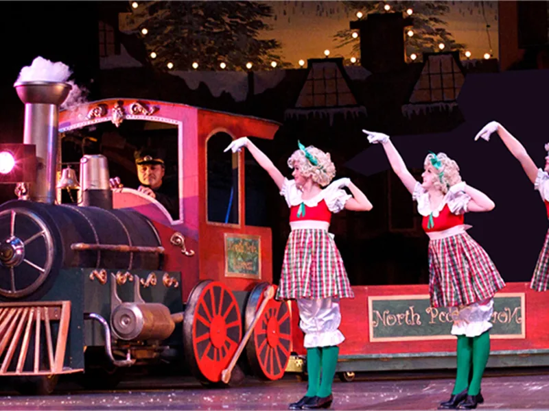 Dancers in plaid dresses and green tights posing beside a red North Pole train.