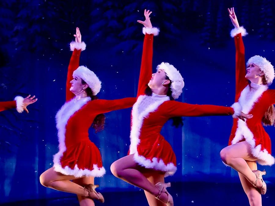 Three dancers in red Santa dresses and white fur hats kicking in unison under blue light.