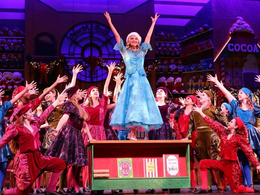 The full cast in Santa's toy shop, arms raised around a girl in a blue dress standing on a toy crate.