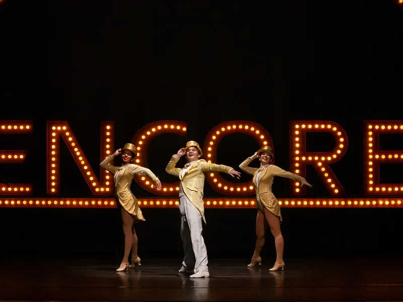 Dancers in gold tailcoats and top hats pose under a glowing ENCORE marquee